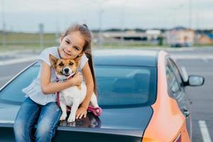 Small attractive female child embraces her favourite dog, sit together at trunk of car, have rest after stroll, enjoy summer day, have friendly relationship. Children, pets and lifestyle concept. photo