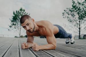 Motivational bearded man stands in plank pose, trains muscles and has strong body. Sporty adult guy performs push up exercise outdoor. Bodybuilder has abdominal workout. Active lifestyle concept photo