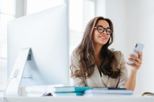 Smiling female in corporate clothes watches funny video via smartphone, sits at desktop in office, uses computer for work, enjoys working process, browses information for project, wears eyeglasses photo