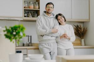 Shot of relaxed family couple stand in kitchen, drink takeaway coffee, wear casual outfit, pose in kitchen interior, bought new apartment for living. People, real estate and mortgage concept photo