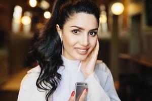 Close up portrait of attractive brunette woman with wavy pony tail, dark eyes and sensual lips having earphones in ears using app to connect with friends on social media. Lifestyle and fashion concept photo