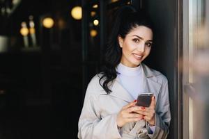 Smiling pretty brunette woman with pony tail having red manicure dressed formally holding smartphone doing online shopping while waiting for train. Businesswoman with cell phone going for work photo