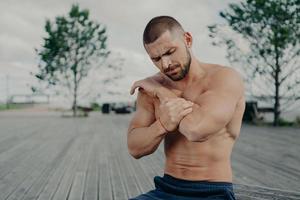 Outdoor shot of muscular guy injured elbow during physical exercises, feels strong pain and poses with naked torso. Health problems, medical, sickness and sport concept. Pain relief. Muscle injury photo