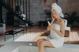 Pleased healthy woman applies body lotion and face cream, nourishes skin, wrapped in bath towel, smiles gently, poses against domestic interior, undergoes beauty treatments. Dermatology concept photo