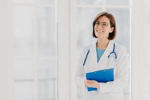 Experienced woman pediatrician stands with clipboard in cabinet, wears white medical coat with stethoscope, gives excellent medical treatment, has happy expression, ready to give consultancy photo