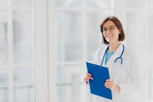 Female cardio therapeutist wears round glasses, white medical coat and phonendoscope, writes with pen in clipboard, stands near big window in own clinic, thinks over patient case and treatment photo