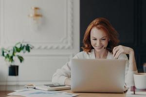 Positive happy female freelancer has busy working day, works distantly from home, sits in front of laptop computer against modern interior, works on creative task, watches webinar for improving skills photo