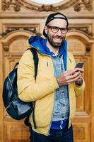 Hipster tourist in glasses, cap and yellow anorak holding backpack and smartphone having excursion in art gallery making photos being happy and delightful. People, tourism, technology concept