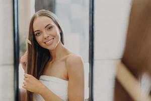 Portrait of beautiful gorgeous woman with bare shoulders combes her straight hair, smiles broadly and looks at her reflection in mirror, poses in bathroom, wrapped in towel. Women beauty concept photo