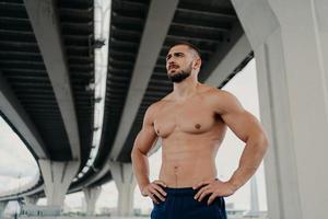 Athletic male fitness model keeps hands on waist poses with muscular perfect naked body, looks seriously into distance, does sport exercises outdoor, focused somewhere, goes in for sport regularly photo