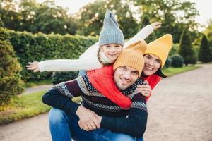 Handsome young man spends his free time with family, recieves embrace from adorable daughter and pretty wife, have picnic during autumn weekend. Happy affectionate family have good relationships. photo