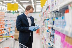Shot of adult man chooses detergent in housekeeping store, reads label and instruction how to use product, wears medical mask and gloves during coronavirus pandemic, avoids risk to catch virus photo