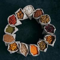 Assorted legumes and beans in sacks standing in circle, isolated over dark background with copy space in middle. Healthy eating concept photo