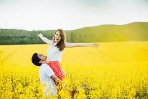 I feel free Joyful young female raises hands being on mans hands, pose together on yellow flower field during sunny summer weather. Romantic couple have fun outdoor. Relationships concept. photo