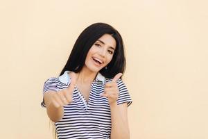 Positive brunette female makes approval gesture, keeps thumbs raised, being in good mood, rejoices something, isolated over light pink background. Optimistic woman agrees or evaluates new project photo