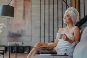 Photo of relaxed smiling woman enjoys drinking tea, reads magazine and poses on comfortable sofa in living room, enjoys spare time during day off, wears minimal makeup, has well cared healthy skin