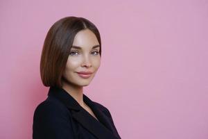 foto de perfil de una mujer de cabello oscuro vestida con traje negro, tiene maquillaje natural, se ve segura de sí misma en el interior, posa sobre fondo rosa, espacio vacío para su promoción. concepto de mujer y elegancia