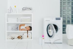una niña cansada descansa en el estante de la consola junto con una mascota, lava en casa, duerme en la lavandería, abre la lavadora con una toalla sucia adentro. infancia, limpieza, concepto de tareas familiares foto