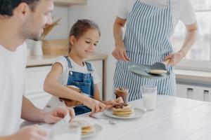 Unrecognizable mommy brings delicious pancakes to table, prepares breakfast for family. Cheerful girl adds melted chocolate to dessert, enjoys time with favourite pet and parents. Breakfast time photo