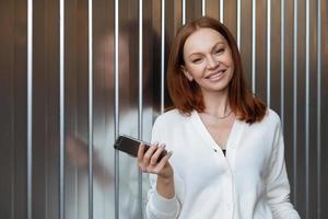 Satisfied female entrepeneur with toothy smile, wears white jumper, holds modern cell phone, sends text message, waits for business partner, stands indoor with free space for your promotion. photo