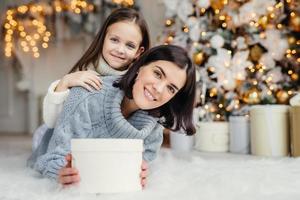 Children, family and celebration concept. Adorable female in knitted sweater holds white present box and small kid stands behind her back, have good mood before New Years Eve. Waiting miracle photo