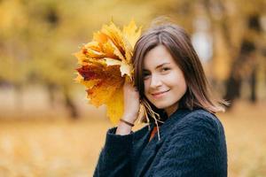 Cute smiling young European woman has stroll in park, enjoys sunny day during autumn, carries yellow leaves, wears coat, poses against blurred background. People, leisure and season concept. photo