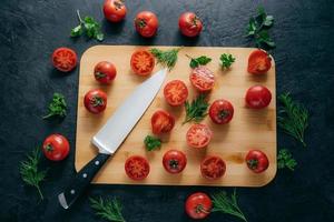 Top view of red sliced tomatoes on wooden chopping board. Sharp knife near. Green parsley and dill. Dark background. Preparing fresh vegetable salad photo