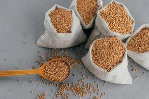 Top view of five small sacks filled with raw buckwheat, wooden spoon near with uncooked cereals, dark background. Healthy food concept photo