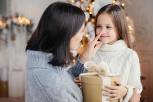 Affectionate mother gives present to her adorble little daughter, prepares surprise on Christmas, touch her nose, expresses great love. Family, celebration, presents, miracle, winter holidays concept photo