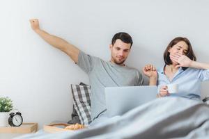 Family spend time together in bedroom, stretch and yawn as get up early, do remote work on laptop computer, use free wifi connection, drink coffee, being busy. Morning and awakening photo
