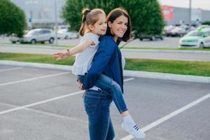 Photo of happy brunette woman dressed in denim clothes, gives piggyback to her small female kid, have positive expressions, poses against city blurred background. Mother, daughter and fun concept