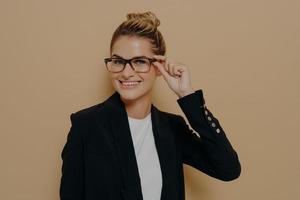 Happy satisfied female teacher in classic black suit smiling broadly and adjusting her glasses with hand photo