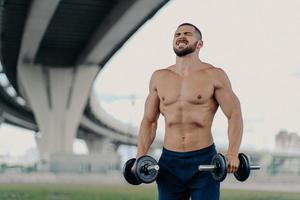 Endurance and power concept. Motivated bearded European man raises barbells puts all efforts in lifting heavy weight clenches teeth poses with sport equipment under bridge, has muscular body photo