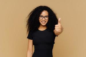 Waist up shot of satisfied supportive woman shows thumb up, cheers best friend, encourages for excellent efforts, wears glasses and black t shirt, stands against beige background. Body language photo