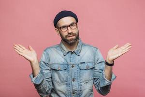 Portrait of hesitant bearded male with doubtful expression, shrugs shoulders, wears denim jacket, glasses and hat, isolated over pink studio background. Confused middle aged man poses indoor photo