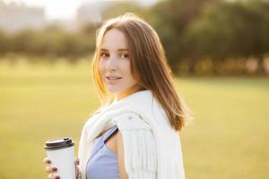 Portrait of good looking young woman with appealing look, enjoys aromatic coffee from disposable cup, has stroll with boyfriend in field, looks happily at camera. People and recreation concept photo