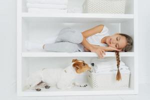 Sleepy little girl feels comfortable as lies on white shelf dressed in casual clothing tired after washing her dog looks attentively. White color. Kid rests in laundry room, enjoys domestic atmosphere photo