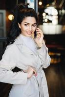 Beautiful young woman with dark hair tied in pony tail and nice eyes dressed in white raincoat speaking over smartphone holding hand on waist while standing outside looking down having happy look photo