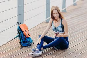 Beautiful serious female has gaze at screen of smart phone, checks which song to listen, uses modern earphones for entertainment, sits crossed legs near bag outdoor, has rest after training. photo