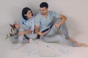 Cheerful young woman uses banking app on her mobile phone, his husband shows figures on calculator, surrounded with papers, have much work, solve economic problems, discuss something together photo