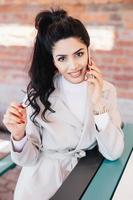 Young charismatic woman with dark wavy pony tail having red manicure dressed eleganltly talking on her mobile phone with smile while sitting at cafe waiting for her order. Communication concept photo