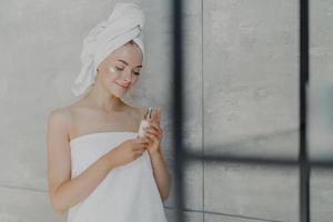 Satisfied young Caucasian woman cares of her beauty, applies lotion cream on face, stands wrapped in bath towel, poses in home bathroom against grey wall. Daily home care and day spa concept photo