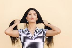Lovely young Caucasian female shows her long straight dark hair, pouts lips, demonstrates natural beauty, poses against light background, dressed in striped t shirt. Hair care and beauty concept photo