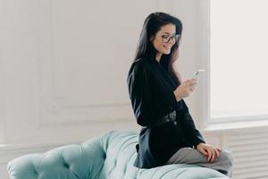 European entrepreneur wears corporate suit and spectacles, uses online banking application for transferring money, holds cellular, sends email, poses at back of comfortable sofa, uses free wifi photo