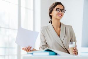 Cheerful businesswoman reads paper documents, focused aside with smile drinks milkshake sits at desktop makes business plan for project. Female executive manager in spectacles poses in coworking space photo
