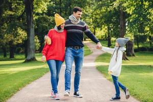 Outdoor portrait of affectionate family walk in park, wear warm knitted clothes. Handsome young man holds daughters hand, looks with happy expression at her. Rest and relaxation concept photo