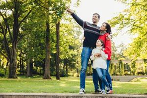 Image of happy joyful young family father, mother and little daughter play together in autumn park, countryside, enjoy nature outside. Attractive man shows something with hand daughter and wife
