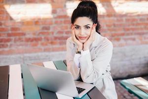 Lovely young brunette woman with charming face and red manicure wearing white coat sitting at table using laptop computer chatting with her family using free internet connection with high speed photo