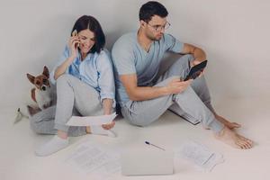 Young woman and man sit back to each other, do accounts together, make necessary calculations, surrounded with paper documents, sit on floor in empty room with dog. People and finances concept photo