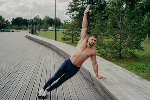 Athletic man stands in side plank pose, raises one arm, poses with shirtless torso, wears trousers and sneaks. Sportsman exercises in park on fresh air, has perfect fit muscles body. Healthy lifestyle